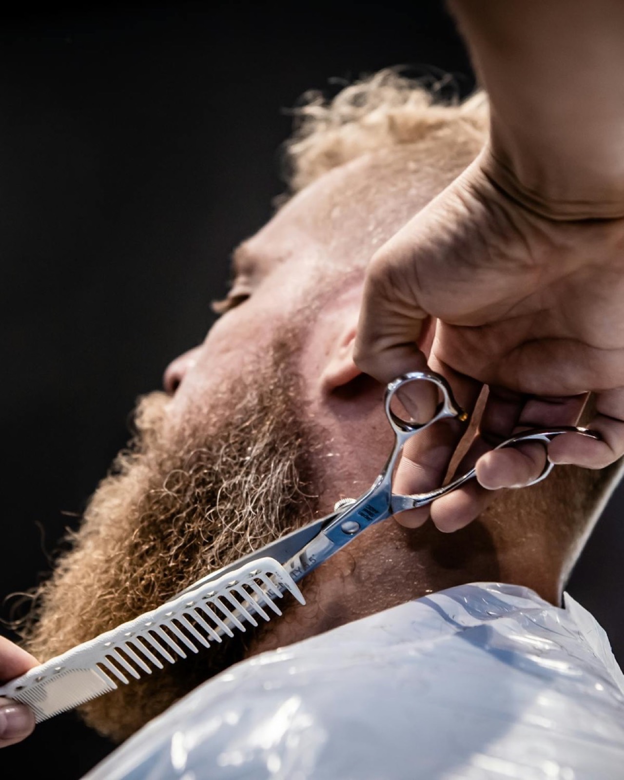 Detail of a barber using shears to shape a ginger beard against a dark background.